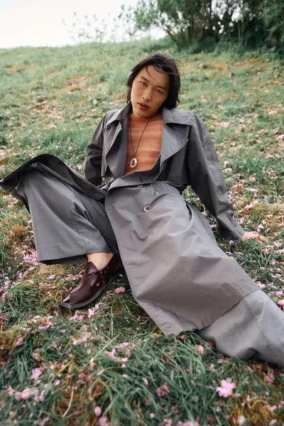 A young Asian man in a grey trench coat sits gracefully in a park, enjoying nature. — Stock Photo