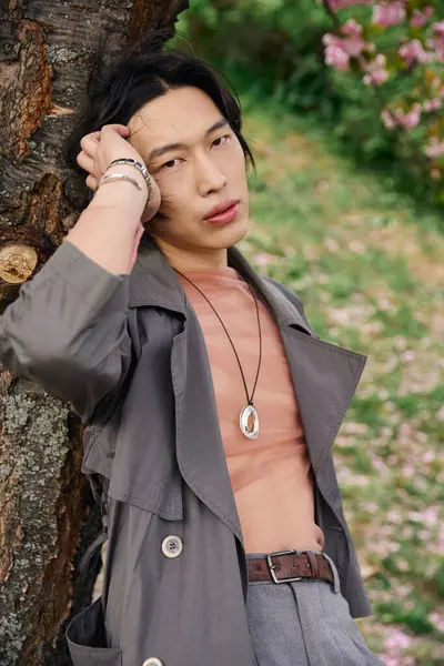 A young asian man poses confidently in a stylish grey outfit, relaxed in a beautiful park setting. — Stock Photo