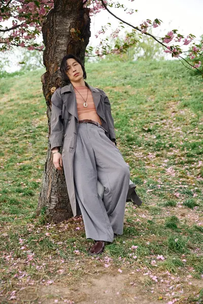 A young asian man stands relaxed against a tree in a park, surrounded by blooming flowers. — Stock Photo