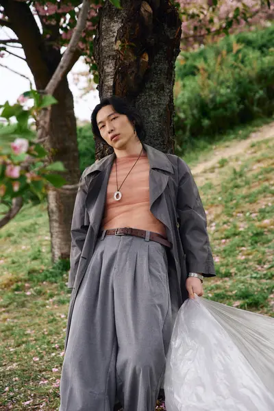 Young man in a grey trench coat leans against a tree, embracing the calm atmosphere in the park. — Stock Photo