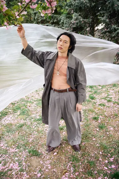 Un joven asiático posa elegantemente en un traje gris de moda sosteniendo polietileno en medio de flores de cerezo florecientes. - foto de stock
