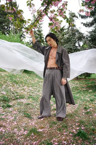 A young Asian man in a fashionable grey outfit stands near polyethylene in a park filled with flowers. — Stock Photo