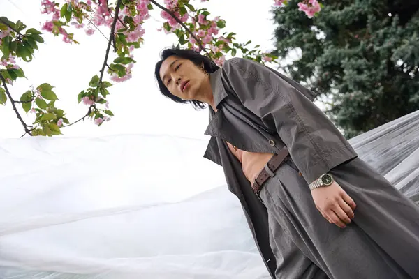 Un jeune homme asiatique modèle une tenue grise élégante dans un parc serein avec des fleurs de cerisier. — Stock Photo