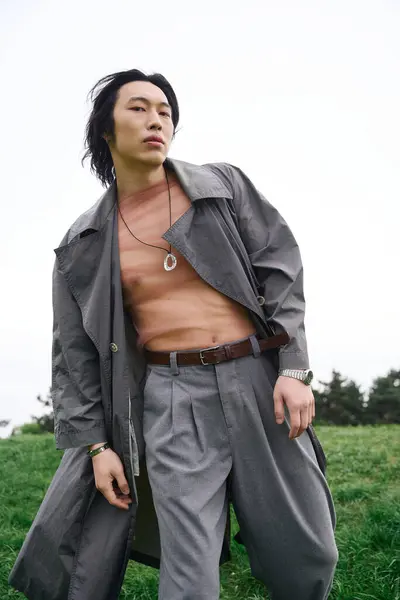A young Asian man in a grey trench coat stands confidently in the park, showcasing his style. — Stock Photo
