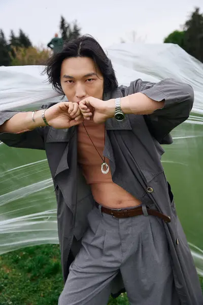 A young asian man confidently poses in a trendy grey outfit in a lush park near polyethylene — Stock Photo