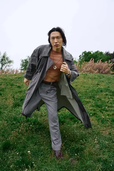 A young asian man in a fashionable grey trench coat strides confidently across the park grass. — Stock Photo