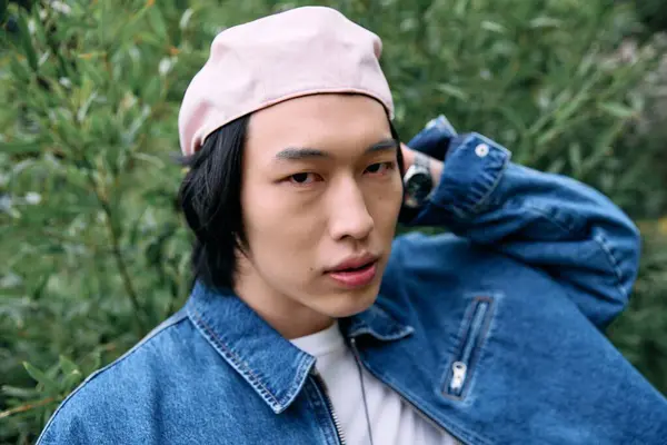 Ein junger asiatischer Mann präsentiert sein modisches Jeans-Outfit, während er sich in einem üppigen Park entspannt. — Stockfoto