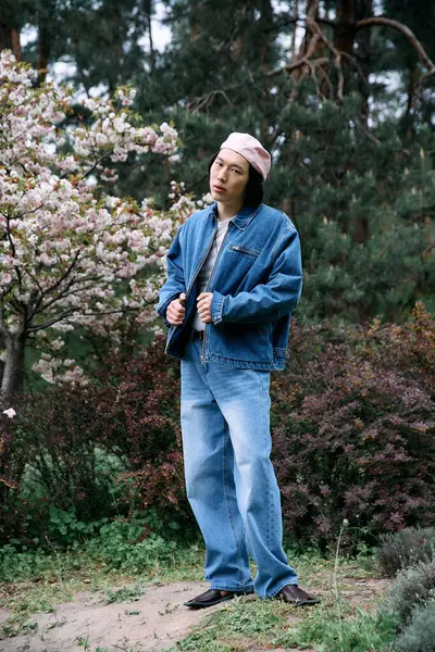 A young asian man stands confidently in a stylish denim outfit amidst blossoming trees in a park. — Stock Photo