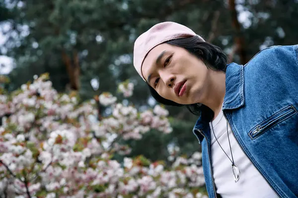 Young asian man wearing a denim outfit stands amidst blooming flowers in the park, enjoying nature. — Stock Photo