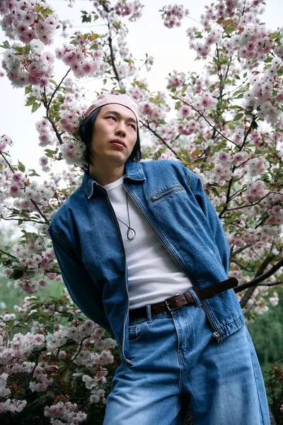 A young Asian man in a trendy denim outfit stands among blooming cherry blossoms, exuding style. — Stock Photo