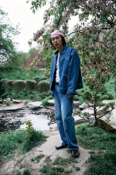 A young man in a chic denim outfit stands confidently in a flower-filled park. — Stock Photo