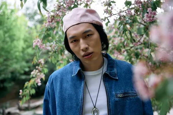 A young asian man poses confidently in a trendy denim outfit surrounded by pink blossoms. — Stock Photo