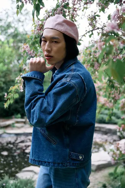 A stylish young man poses confidently in a denim outfit among blooming trees in the park. — Stock Photo