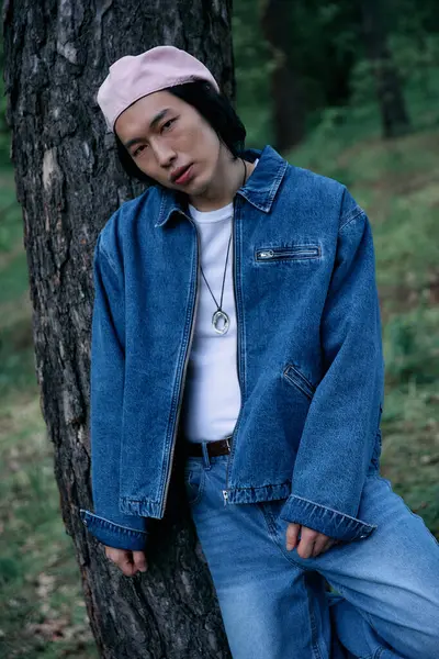 A young asian man wearing denim stands by a tree, radiating style and confidence in a serene park. — Stock Photo