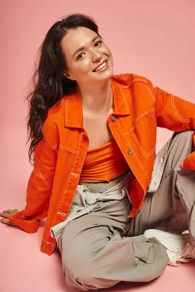A joyful young woman relaxes in a trendy orange outfit against a bright pink background. — Stock Photo