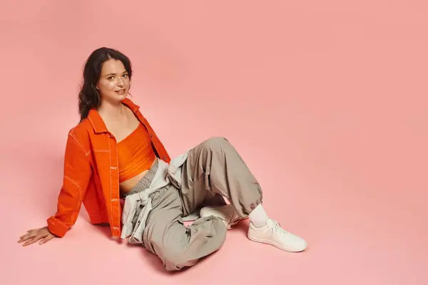 A young woman sits casually in trendy attire, radiating confidence against a pink backdrop. — стоковое фото