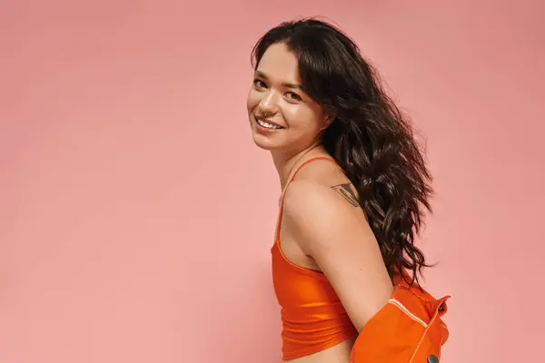 A young woman showcases her fashionable orange outfit, smiling joyfully in front of a pink backdrop. — Stock Photo