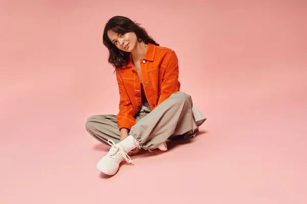 Young woman exudes confidence in a striking orange jacket, sitting gracefully on a pink surface. — Stock Photo