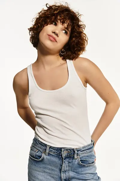 Una mujer joven con el pelo rizado posa con confianza en una camiseta sin mangas y pantalones vaqueros sobre un fondo brillante. — Stock Photo