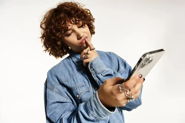 Mujer joven con el pelo rizado cuidadosamente se involucra con su teléfono inteligente mientras está vestida con denim casual. — Stock Photo
