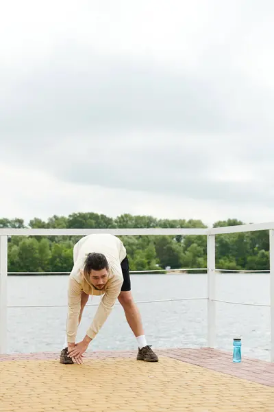Young man in a hoodie is stretching by the water outdoors in a serene setting. — Stock Photo