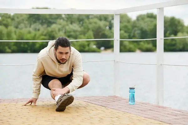 Ajuste joven en una sudadera con capucha se extiende mientras hace ejercicio cerca del agua, disfrutando de la naturaleza. - foto de stock