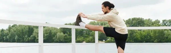 A young athletic man in a hoodie performs a stretch by the water, enjoying his workout routine, banner — Stock Photo