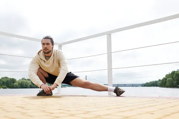 Sporty individual wearing a hoodie practices stretches by the water in an outdoor setting. — Stock Photo
