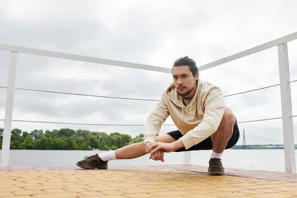 Enfocado joven en una sudadera con capucha se involucra en estiramiento al aire libre, disfrutando del aire fresco junto al agua. - foto de stock