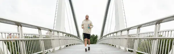Un joven atlético en una sudadera con capucha corre enérgicamente a través de un puente moderno, abrazando la aptitud. - foto de stock