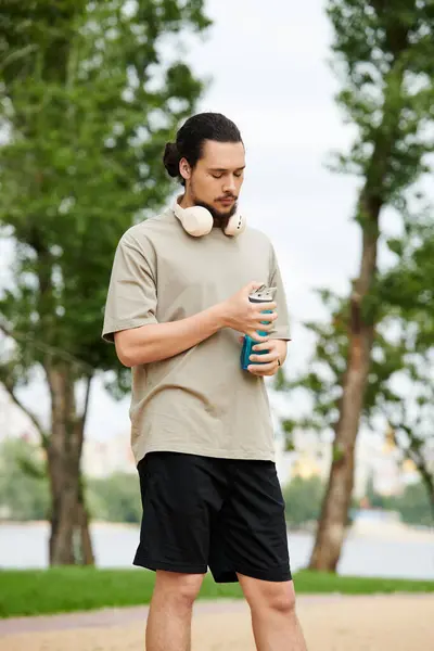 Sporty young man rests outdoors after workout, sipping a drink amidst lush greenery and sunshine. — Stock Photo