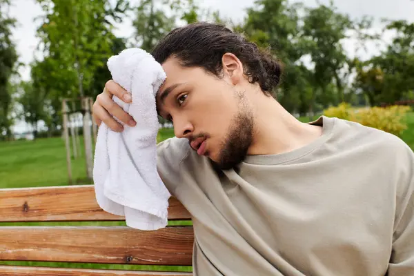 Un joven se limpia el sudor de la frente mientras descansa en un banco después de un duro entrenamiento al aire libre. - foto de stock