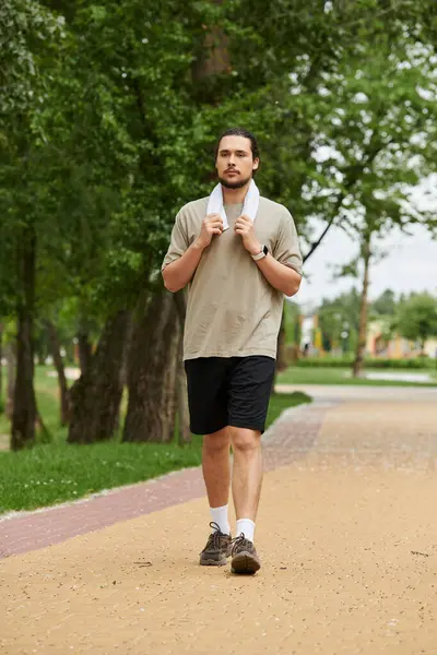 A bearded young man walks along a path in a green park, towel around his neck, exercising. — Stock Photo