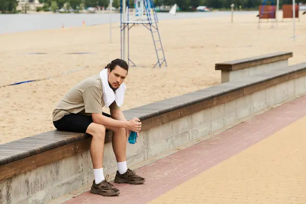 A young man rests on a bench, enjoying a drink after exercising outdoors near the beach. — Stock Photo