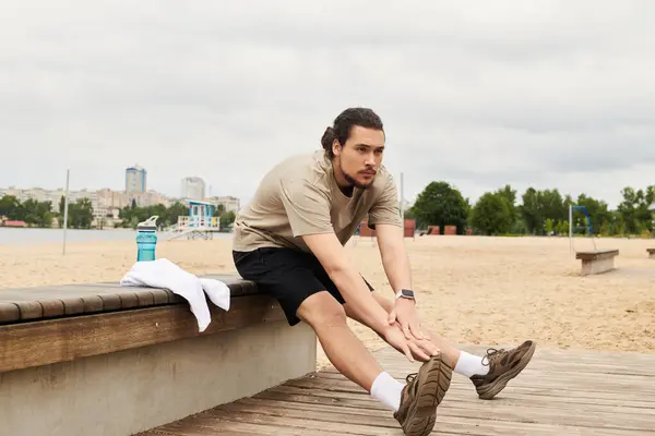 A fit young man with a beard is stretching on a bench at an outdoor beach, focusing on his workout. — Stock Photo