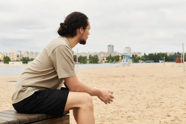 Fit young man with a beard sits thoughtfully on the sand after exercising outdoors. — Stock Photo