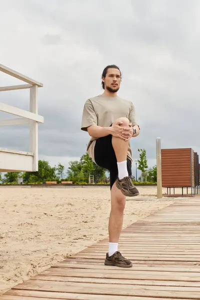 A young man with a beard is doing a leg stretch on a wooden path by the beach. — Stock Photo