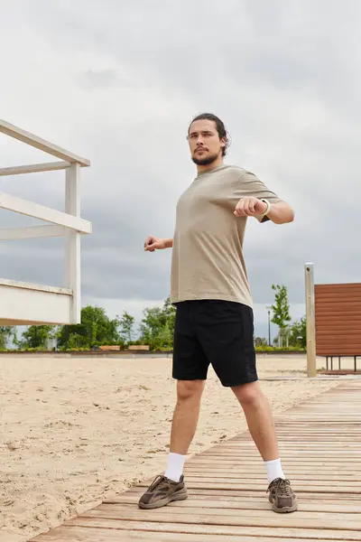 Young man stretches and prepares for a workout outdoors on a sandy beach boardwalk. — Stock Photo