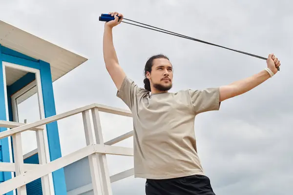 A young man with a beard stretches with a resistance band outside near a lifeguard tower. — Stock Photo