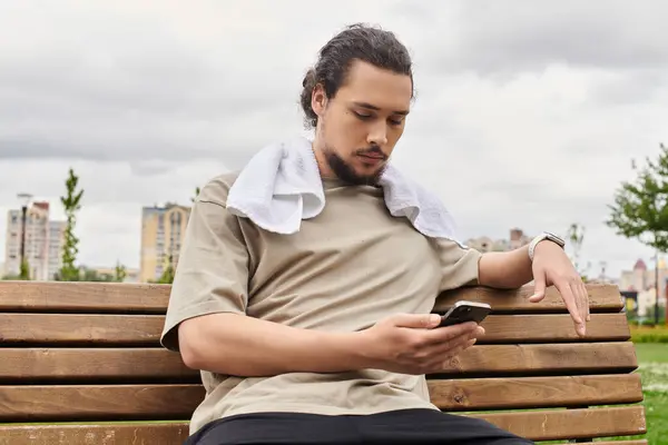 A fit man sits on a bench, resting after his workout and looking at his phone in a park. — Stock Photo