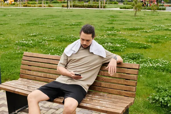 A sporty young man takes a moment to rest on a bench, checking his phone outside in nature. — Stock Photo