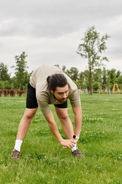 Joven con barba realiza un ejercicio de estiramiento en un parque durante un día nublado. - foto de stock