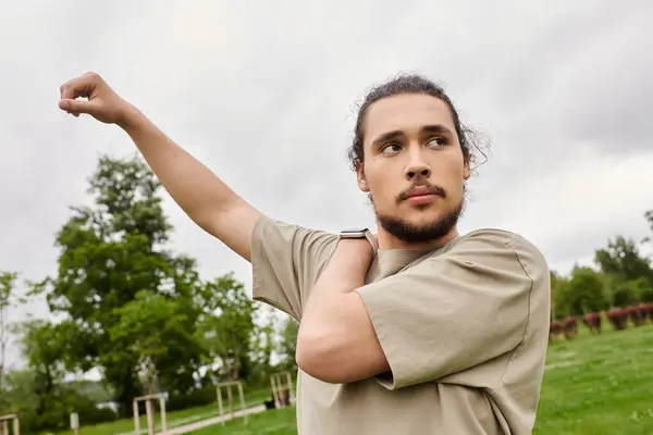 A young man with a beard performs a stretching exercise in a green outdoor park setting. — Stock Photo