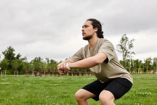 A fit young man with a beard performs squats outdoors, enjoying a cloudy day in nature. — Stock Photo