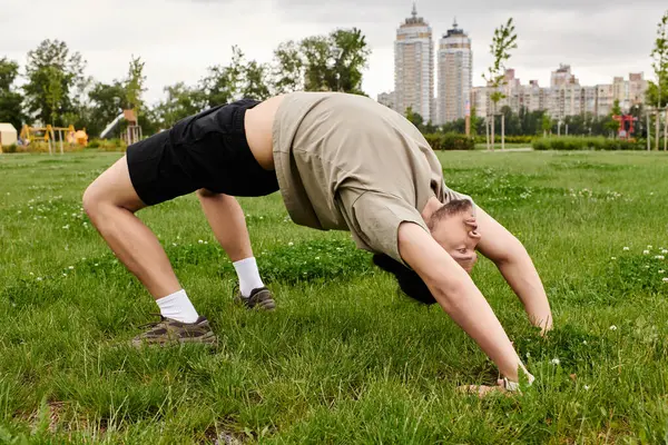 A bearded young man stretches impressively in a green park near buildings. — Stock Photo