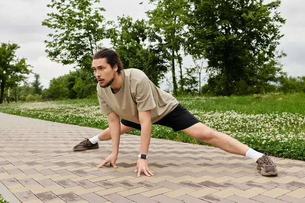 A bearded young man stretches on a paved path, surrounded by greenery, enjoying exercise. — Stock Photo