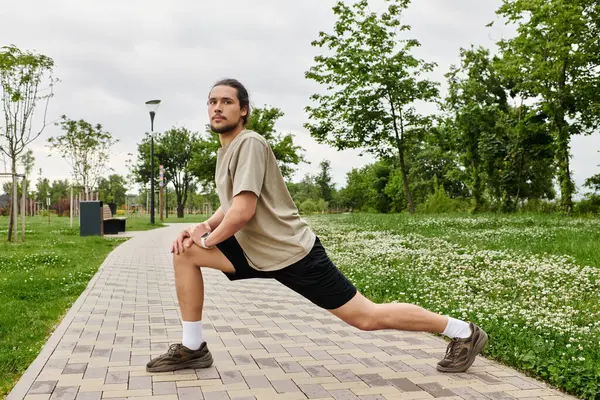 Sporty man with beard stretches by the path in a green park to boost his athletic performance. — Stock Photo