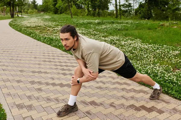 A young man with a beard stretches on a walkway surrounded by lush greenery during the day. — Stock Photo