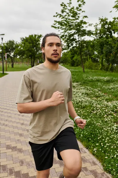 A fit young man with a beard is jogging energetically in a green park on a cloudy day. — Stock Photo