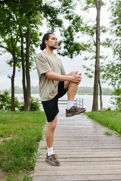 A young, fit man with a beard performs an exercise outdoors, enjoying nature by the lake. — Stock Photo
